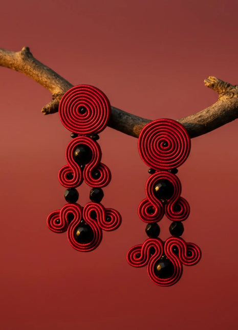 Pendientes Versalles de soutache rojo con cristales negros hechos a mano en Bizkaia, colgando de rama sobre fondo rojo