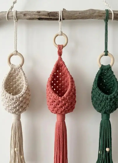 Tres maceteros colgantes de macramé Gota tejidos a mano en colores natural, rosa y verde oscuro colgados de una rama de madera