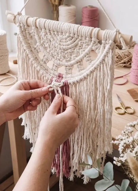 Manos tejiendo el tapiz de macramé Calma en taller con carretes de cuerda natural y rosa — kit de manualidades, regalo original para mujer